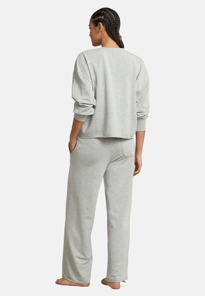 Γυναίκα με πλεξούδες στέκεται ξυπόλητη, με την πλάτη γυρισμένη, φοράει γκρι ανοιχτό γκρι μακρυμάνικη φούτερ μπλούζα και αντίστοιχα φαρδιά φόρμα με τα χέρια στις τσέπες.