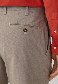 Pantalones de cuadro en tono beige con un bolsillo trasero que cuenta con cierre de botón. La tela parece suave con una textura sutil. Combinados con una camisa roja.