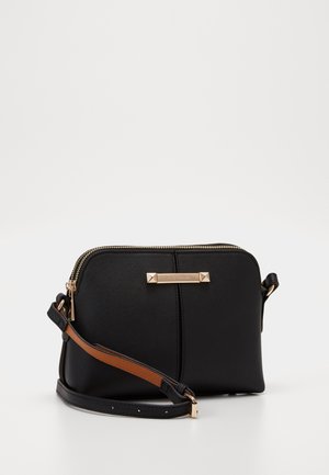 Petit sac bandoulière noir avec finition texturée, fermeture éclair dorée et sangle marron ajustable, présenté sur un fond blanc uni.