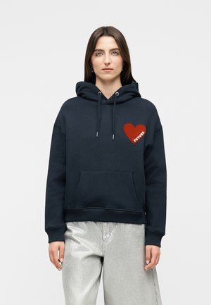 Naine, kes kannab tumel sinist kapuutsiga pusa punase südame ja sõnaga "FUTURE" rinnal, kombineeritud heledate hallide pükstega.