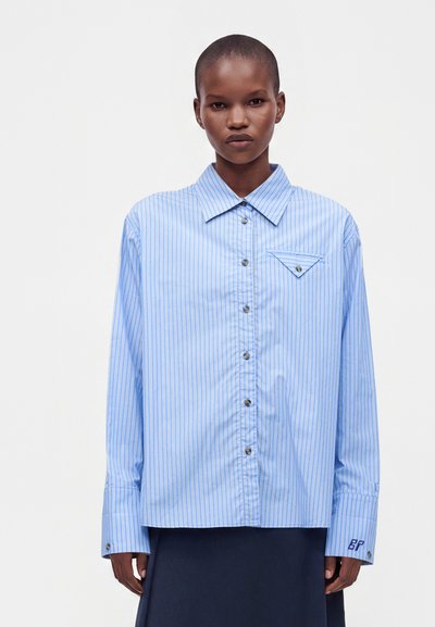 Personne portant une chemise oversize rayée bleu clair et blanc avec une petite poche sur la poitrine et des initiales sur la manche.
