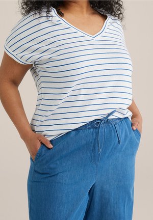 Kobieta w białej koszulce z dekoltem w serek z niebieskimi poziomymi paskami i niebieskich spodniach z sznurkiem, ręce w kieszeniach, na jednolitym tle.