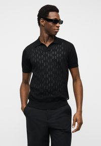 Νέος άνδρας που φοράει μαύρο πουκάμισο πόλο με σχέδιο και κοντά μανίκια, μαύρο παντελόνι και λεπτά μαύρα γυαλιά ηλίου, στέκεται με το ένα χέρι στην τσέπη.