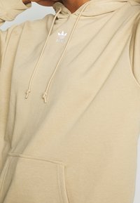 Sudadera con capucha beige de tela suave, con un bolsillo frontal, cordones y un pequeño logo blanco de Adidas en el pecho.