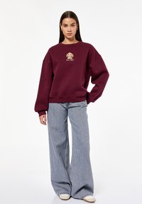 Bordowa bluza z okrągłym dekoltem i opadającymi ramionami, z grafiką psa na piersi. W komplecie ze luźnymi szarymi dżinsami.
