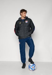 Svart boblejakke med røde og blå detaljer, med en logo. Kombinert med marineblå joggebukser. Spilleren står på en flerfarget fotball.
