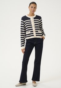 Navy and cream striped cardigan, buttoned front, with pockets. Paired with dark denim flared pants and light-colored shoes.