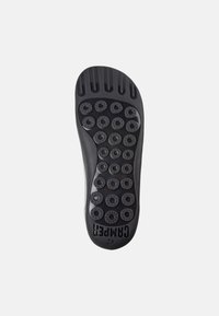 Suola di calzature nera con grip in gomma circolare, scanalature testurizzate e branding. Presenta punti in rilievo per una maggiore trazione e flessibilità.
