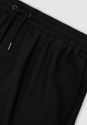 Černé krátké kalhoty z texturované tkaniny s elastickým pasem a černým šňůrkovým stahováním s kovovými zakončeními na světlém pozadí.