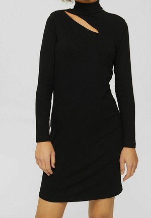 Femme portant une robe noire à manches longues en maille côtelée avec un col montant et une découpe asymétrique près de l'épaule droite.