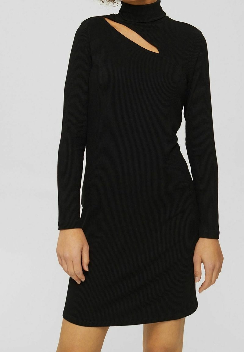 Femme portant une robe noire à manches longues en maille côtelée avec un col montant et une découpe asymétrique près de l'épaule droite.