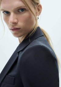 Mustat räätälöidyt bleiserit, joissa on rakenteelliset olkapäät, sujuva kangas ja yksi rintatasku. Mallilla on pitkä hiusstyylit taakse, mikä tuo esiin hopeiset korvakorut.