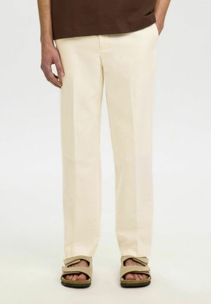 Osoba oblečená v krémové rovné nohavice a béžové sandále s otvorenou špičkou stojí na jednotnom pozadí.