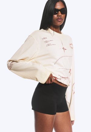 Femme portant un sweat-shirt crème oversize avec des motifs rouges abstraits et un short noir moulant, posant avec une main sur la hanche, portant des lunettes de soleil noires.