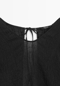 Sort tekstureret stoftop med rundet halsudskæring og bindelukning bagpå, med et lille mærkeindvendigt mærke i kraven.