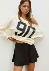 Kremast sweatshirt oversized kroja s crnim printom "90", u kombinaciji s crnom mini suknjom na zvono. Glatka tkanina, ležeran fit.