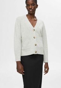 Šviesiai pilkas vilnonis kardiganas su V formos iškirpte, penkiais rudi mygtukais, raukšlėmis rankovėse ir dviem priekinėmis kišenėmis, derinamas su juoda sijonu.