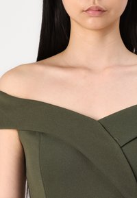 Abito off-shoulder verde oliva con un design strutturato, caratterizzato da un corpetto incrociato e una texture liscia. Lo scollo mette in risalto le clavicole.
