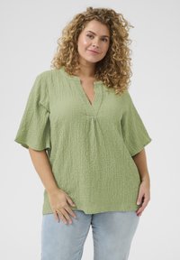 Light green, textured blouse with a V-neck, short flutter sleeves, and subtle gathering at the front. Paired with light blue jeans.