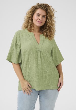 Lichtgroene, gestructureerde blouse met een V-hals, korte flutter mouwen en subtiele plooien aan de voorkant. Combinatie met lichtblauwe jeans.