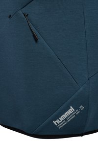 Blauwgroene stof met diagonale naden, een zwarte koordtrekker voor de rits, en een wit "hummel" sportkledinglogo en tekst op de onderzoom.