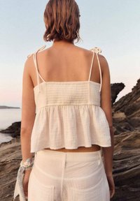 Bílý, strukturovaný top na ramínka s nastavitelními šňůrkami, peplum lem a odpovídající lehké kalhoty, zasazené do pobřežního prostředí.