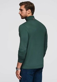 Grünes Langarmshirt mit hohem Kragen, mit einem kleinen roten Akzent auf der Rückseite. Glatter Stoff, taillierter Schnitt, kombiniert mit blauen Jeans.