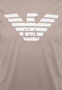 Ljusbrun T-shirt i mjukt tyg som har en stor vit örnlogotyp med parallella linjer, tillsammans med "GA" i matchande färg under emblemet.