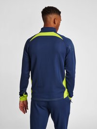 Marinblå idrottsjacka med neongröna detaljer, som har en dragkedja vid kragen, texturerade axlar och meshdetaljer på sidorna.