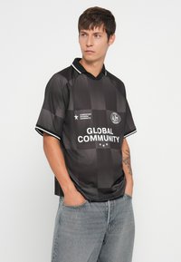 Νεαρός άνδρας με κοντά καστανά μαλλιά, φορώντας μαύρο καρό πουκάμισο με την επιγραφή "ΠΑΓΚΟΣΜΙΑ ΚΟΙΝΟΤΗΤΑ", στέκεται με τα χέρια στις τσέπες σε λευκό φόντο.