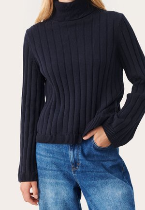 Woman wearing a navy ribbed knit turtleneck sweater and blue denim jeans, with one hand in her pocket.
