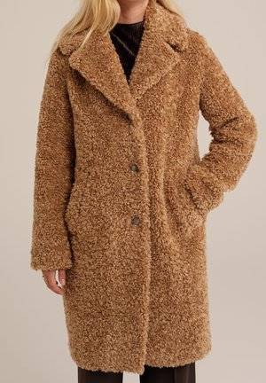 Femme portant un manteau en fausse fourrure bouclée de longueur moyenne, couleur beige, avec de grands revers et une fermeture à boutons, main dans la poche, sur un fond uni.