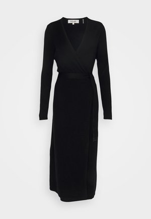 Kobieta siedząca na białym fotelu, ubrana w czarną sukienkę z długim rękawem oraz ciemnobrązowe, wysokie kozaki na obcasie z zaostrzonym noskiem.