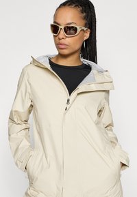 Veste légère beige avec capuche, fermeture éclair à l'avant et poches latérales. Portée sur une chemise noire, assortie de lunettes de soleil marron.