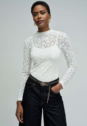 Femme portant un haut à manches longues en dentelle blanche à motifs floraux, rentré dans un jean foncé avec une ceinture chaîne dorée, debout devant un fond gris uni.