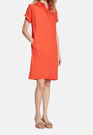 Femme portant une robe chemise orange, manches courtes, longueur genou, avec col et boutons, ainsi que des sandales marron tressées à talons.
