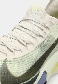 Αθλητικό παπούτσι Nike με πλεκτό επάνω μέρος σε λευκό και γκρι χρώμα με υφασμάτινες λεπτομέρειες, πράσινη λεπτομέρεια και μαξιλαρωτή σόλα.