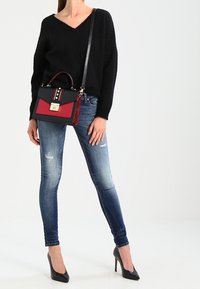 Bolso de mano negro y rojo con un diseño estructurado, que presenta una solapa delantera, herrajes metálicos y una correa texturizada. Llevado con jeans ajustados y tacones negros.