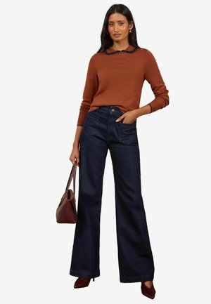 Vrouw staand met een roestkleurige trui met een ruches kraag, donkere wijde jeans, bruine hakken en een bruine handtas in haar hand.