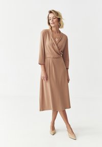 Beige kääritav kleit, millel on kolmveerandvarrukad ja midi pikkus, pehme kangas ja ümar kaelus. Kandmine beežide kortsudega ja lihtsa ehega.