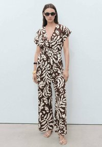 Jumpsuit s uzorkom u smešnim i krem nijansama, kratkih rukava, s vezivanjem u struku, širokim nogavicama i dubokim V-izrezom. Model nosi crne naočale i sandale.