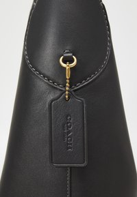 Sac à main Coach en cuir noir avec bords cousus et étiquette en cuir noir embossée « COACH New York », attachée par une chaîne à billes dorée.