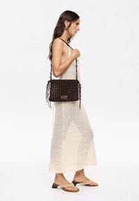 Borsa a tracolla in tessuto marrone con forma rettangolare, frange decorative e superficie testurizzata. Cinghia regolabile e un dettaglio di branding sottile.