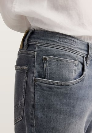 Nahaufnahme einer Person, die verblasste blaue Jeans mit Vorder- und Gesäßtaschen trägt, kombiniert mit einem in die Hose gesteckten weißen Hemd.
