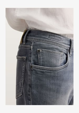 Nahaufnahme einer Person, die verblasste blaue Jeans mit Vorder- und Gesäßtaschen trägt, kombiniert mit einem in die Hose gesteckten weißen Hemd.