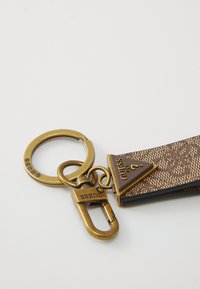 Porte-clés en tissu marron à motifs avec des anneaux en laiton, un fermoir et un accent en forme de triangle portant le logo Guess sur un fond blanc.