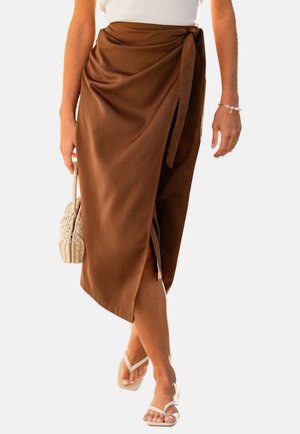Donna che indossa una gonna a portafoglio marrone con uno spacco laterale, porta una borsa intrecciata e indossa sandali bianchi e un top bianco.