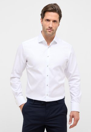 Mand med brunt hår iført en tætsiddende hvid skjorte med knapper og mørke bukser, stående mod en ensfarvet lys baggrund.