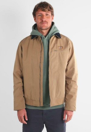 Hombre vistiendo una chaqueta de cremallera color beige sobre una sudadera verde y pantalones oscuros, de pie contra un fondo claro y sencillo.