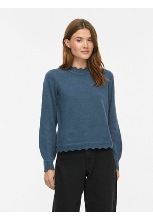 Femme aux longs cheveux bruns portant un pull bleu à manches longues avec des bords ondulés et un pantalon noir, debout devant un fond blanc.